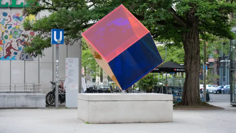 Eine moderne, schräg stehende Skulptur in Würfelform mit pinken und blauen Glasflächen im Außenbereich.