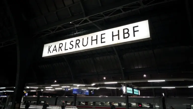 Beleuchtetes Schild des Karlsruher Hauptbahnhofs bei Nacht mit einem einfahrenden Zug im Hintergrund.