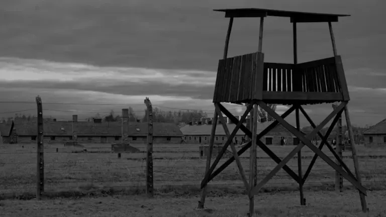 Wachturm des Konzentrationslagers Auschwitz-Birkenau unter einem dramatischen, bewölkten Himmel in Schwarz-Weiß.