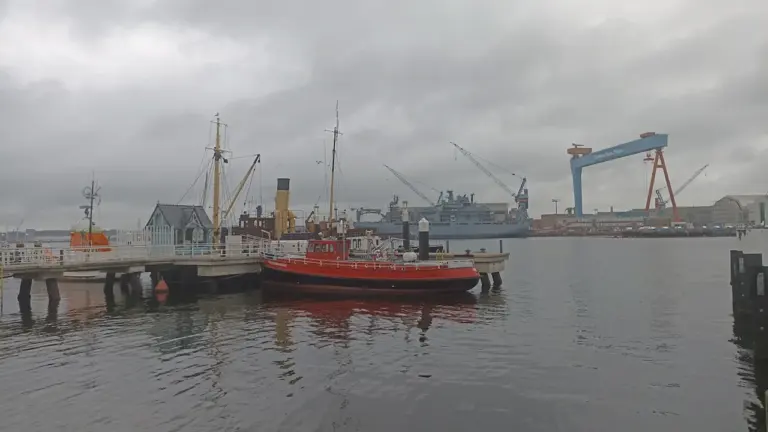 Ein rot-weißes Schiff liegt im Kieler Hafen vor industrieller Kulisse bei bewölktem Himmel.